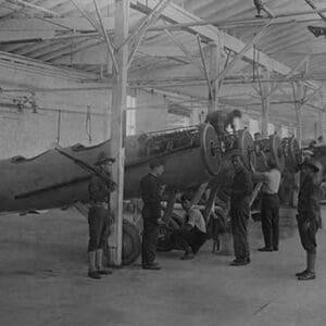 A line of airplanes is being assembled under the watchful eye of a gun-toting soldier as they are being completed to be sent to the European war; the factory is at College Point